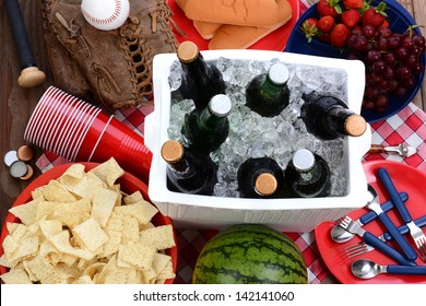 Overhead View Of A Picnic Table, With Ice Chest Full Of Beer, Bowl Of Chips, Watermelon, Strawberries, Grapes, Corn Chips, Plastic Cups, Hot Dog Buns And Baseball Equipment.