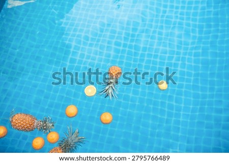 Overhead view of pineapples and oranges floating in a bright blue swimming pool, creating a playful tropical summer pattern.