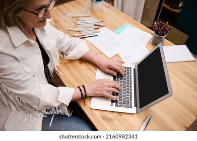 Overhead View Of A Pleasant European Senior Woman Dressmaker Typing Text On Laptop Keyboard Sitting At A Wooden Desk In Fashion Design Atelier. Tailoring Scissors And Fashion Sketches On The Table