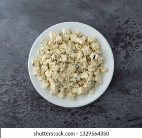 Overhead View Of A Portion Of Crumbled Blue Cheese On A White Plate Atop A Gray Background Illuminated With Natural Lighting.