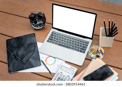 Overhead view of a productive workspace featuring a laptop with blank screen mockup, alarm clock, tablet, glasses, calculator, and colorful stationery on a wooden desk.