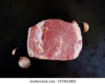 Overhead View Of Raw Piece Of Pork With Fresh Garlic Isolated On Black Background. Piece Of Fresh Boneless Pork, Neck Part Or Collar. Big Piece Of Red Raw Meat On A Black Background. Food Concept.
