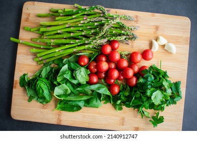 Overhead View Of Raw Vegetables, Herbs, And Spices On A Wooden Cutting Board: Cherry Tomatoes, Asparagus, Basil, Mint, Parsley, And Garlic Cloves On A Bamboo Chopping Board