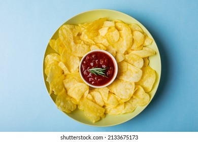 Overhead View Of Red Sauce In Bowl Amidst Potato Chips Served In Plate On Blue Background. Unaltered, Unhealthy Food, Snack, Dipping Sauce, Herb, Crunchy, Salty Snack And Savory Food.