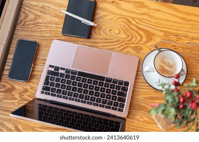 An overhead view of a remote work setup featuring a laptop, smartphone, notebook, and a cup of coffee on a wooden cafe table, accented with a decorative plant.