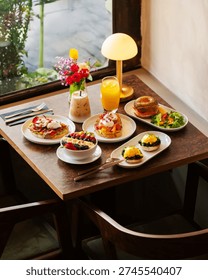 An overhead view of a rich brunch table featuring various avocado toasts, eggs benedict, a fruit-filled smoothie bowl, a loaded bagel, orange juice, and iced coffee under warm lamp lighting.