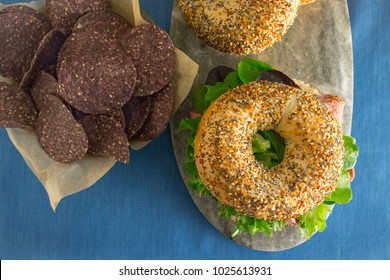 Overhead View Of Roast Beef Sandwich With Lettuce On An Everything Bagel With A Side Of Blue Corn Chips On A Blue Background