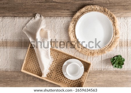 Overhead view of rustic chic inspired table setting featuring white ceramic plate on braided jute charger, woven rattan tray with cream napkin and cup, striped runner and succulent on wood surface.