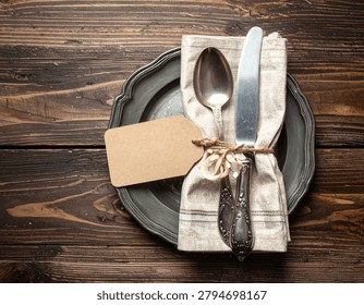 Overhead view of a rustic table setting featuring a vintage pewter plate, cloth napkin, spoon, and knife, with a blank tag for personalization, on a weathered wooden surface.