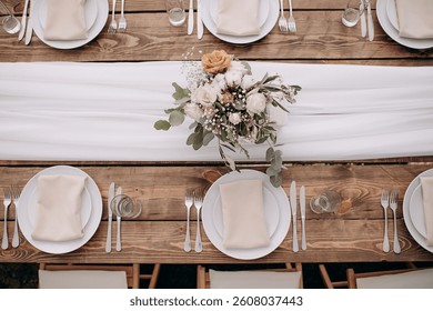 Overhead view of a rustic wedding table setting with a wooden table, white runner, white plates, and a floral centerpiece