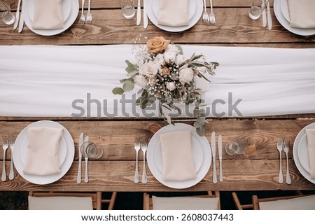 Overhead view of a rustic wedding table setting with a wooden table, white runner, white plates, and a floral centerpiece