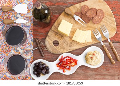 Overhead View Of Savory Snacks On A Rustic Table With Two Glasses And A Bottle Of Red Wine And Havarti Cheese,spicy Dried Sausage,olives, Red Bell Pepper And Hummus With Cheese Cutters For Serving