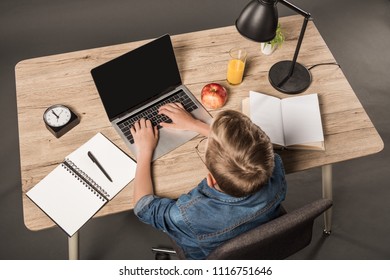 Overhead View Of Schoolboy Doing Homework On Laptop At Table With Textbook, Book, Plant, Lamp, Clock, Apple And Glass Of Juice 