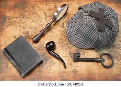 Overhead View Of Sherlock Holmes Deerstalker Hat  And Private Detective Tools On The Old World Map Background. Items Include Vintage Magnifying Glass, Retro Key, Hand Book Or Notepad, Smoking Pipe