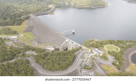 An overhead view showing a large dam embankment holding a reservoir, with roads, trees, and parking areas surrounding the serene water.