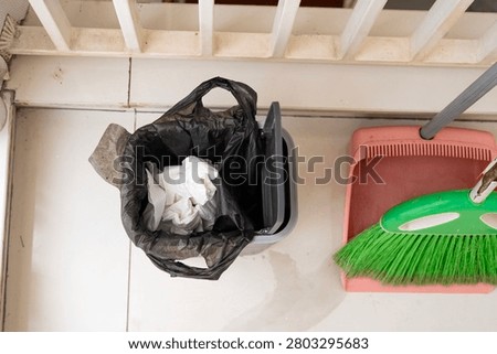 Overhead view of a small wastebasket lined with a black plastic bag containing used white tissues on a tiled floor near a white railing