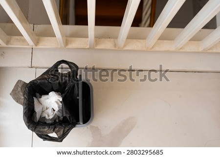 Overhead view of a small wastebasket lined with a black plastic bag containing used white tissues on a tiled floor near a white railing