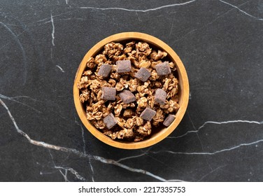 Overhead View Of A Small Wood Bowl Filled With Chocolate Chunk Granola On A Tile Tabletop.