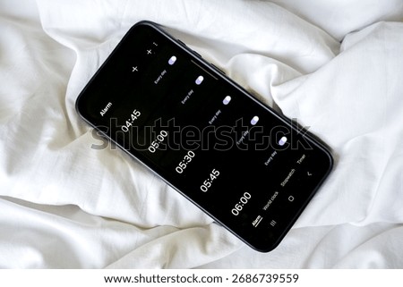 Overhead view of a smartphone displaying multiple alarms on a rumpled white sheet or blanket.