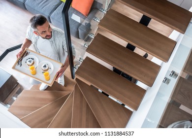 Overhead View Of Smiling Man With Breakfast On Wooden Tray Walking Up Stairs At Home