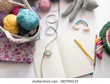 Overhead View Of Some Sewing And Crochet Things On A White Table. In The Left There Is A Crochet Bag Full Of Colorful Yarn Balls, In The Center A White Opened Notebook.