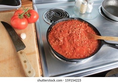 Overhead View Of Spaghetti Sauce In Cast Iron Skillet On Stainless Steel Electric Stove.  Fresh Tomatoes On Cutting Board With Clove Of Garlic And Kitchen Knife.