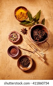 Overhead View Of Spice Selection In Small Jars Besides Dried Cinnamon Sticks And Fresh Leaves On Cracked Paint Background