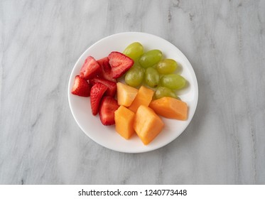 Overhead View Of Strawberries Grapes And Cut Cantaloupe On A White Plate Atop A Gray Marble Counter Top.