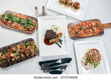 Overhead View Of A Table Full Of A Flatbread Choices, Filet Mignon, Pork Chop, Brussel Sprouts, Or Scallops.