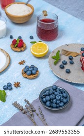 An overhead view of a table with a light blue tablecloth displaying a blueberry cheesecake, alongside two refreshing drinks