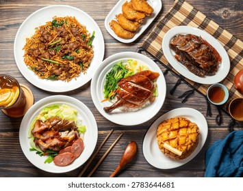 Overhead view of a table with multiple plates of food including noodles, chicken, meat, vegetables, and a pastry, accompanied by chopsticks, a spoon, and small bowls of sauce on a wooden surface