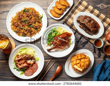 Overhead view of a table with multiple plates of food including noodles, chicken, meat, vegetables, and a pastry, accompanied by chopsticks, a spoon, and small bowls of sauce on a wooden surface