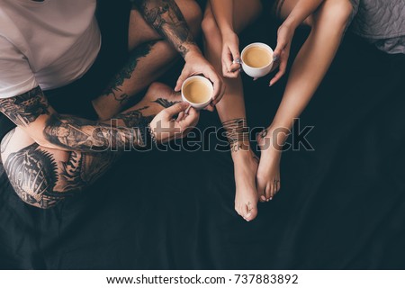 overhead view of tattooed couple with cups of aromatic coffee in bed