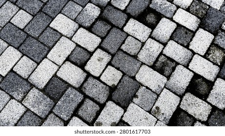 An overhead view of a textured interlocking brick paver walkway featuring a basketweave pattern of grey black and white stone blocks filling the entire frame on a bright day
