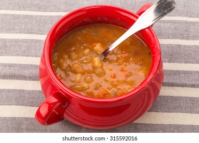 Overhead View Of Thick And Hearty Bean Soup In Red Crock Pot On Gray Striped Tablecloth.