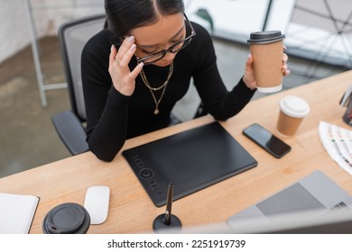 Overhead view of tired asian interior designer holding coffee near devices in studio - Powered by Shutterstock
