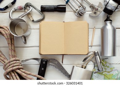 Overhead view of travel equipment for a mountain trip on a rustic wood floor with empty check list in the notebook. Items include flask, map, ice axe, knife, rope, carbine, cup, flashlight 