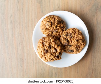 Overhead View Of Two Homemade Oatmeal Raisin Cookies On A White Plate Atop A Table.