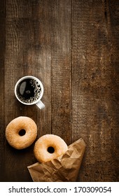 Overhead View Of Two Tempting Fresh Sugared Doughnuts With Their Brown Paper Wrapping And A Cup Of Strong Black Filter Or Espresso Coffee On A Rustic Wood Table