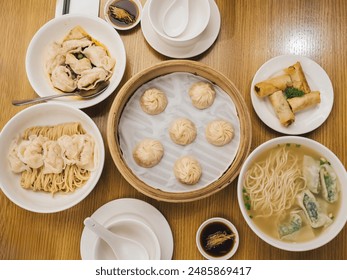 An overhead view of a variety of Chinese dishes, including steamed dumplings in a bamboo basket, noodles with dumplings, spring rolls, and a bowl of dumpling soup, set on a wooden table. Asian Food - Powered by Shutterstock