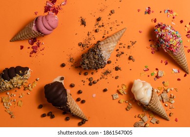 Overhead View Of A Variety Of Soft Serve Ice Cream Cones And Their Toppings On An Orange Background.