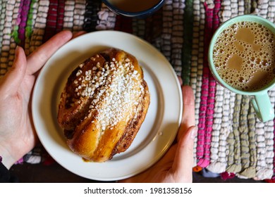 Overhead View Of A Warm Cinnamon Bun On A White Plate With A Fresh Cup Of Coffee On The Side And A Tablecloth With Bright Colors