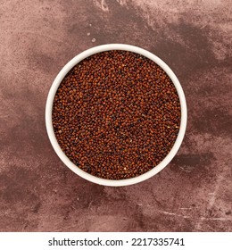 Overhead View Of A White Bowl Filled With Red Quinoa On A Red Mottled Tabletop.