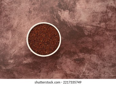 Overhead View Of A White Bowl Filled With Red Quinoa Offset On A Red Mottled Tabletop.