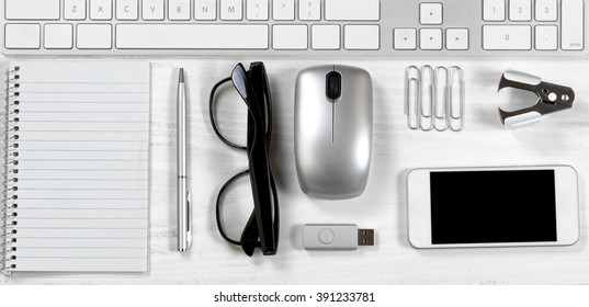 Overhead view of a white desktop with basic office stationery objects lined up under keyboard.