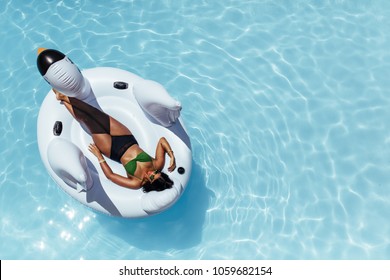 Overhead View Of Woman In Bikini Resting On An Inflatable Swan In Swimming Pool. Female Sunbathing On Inflatable Toy Floating In Pool.