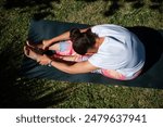 Overhead view of a woman doing a seated forward bend yoga pose on a mat outdoors. She is enjoying a healthy lifestyle.