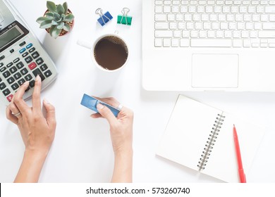 Overhead View Of Woman Hand Using Calculator And Holding Credit Card With Laptop And Stationery On White Table