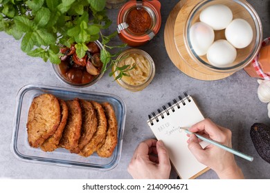 Overhead view of a woman hand writing in a spiral notebook BATCH cooking next to some glass jars full of meal. - Shutterstock ID 2140904671