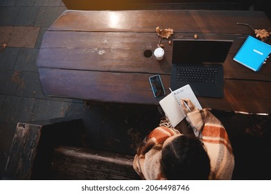 Overhead View Of A Woman Writing In Notebook While Sitting On A Wooden Bench In Front Of A Laptop And Office Supplies. Concept Of E-learning, Remote Work, Remote Business During Quarantine Lockdown